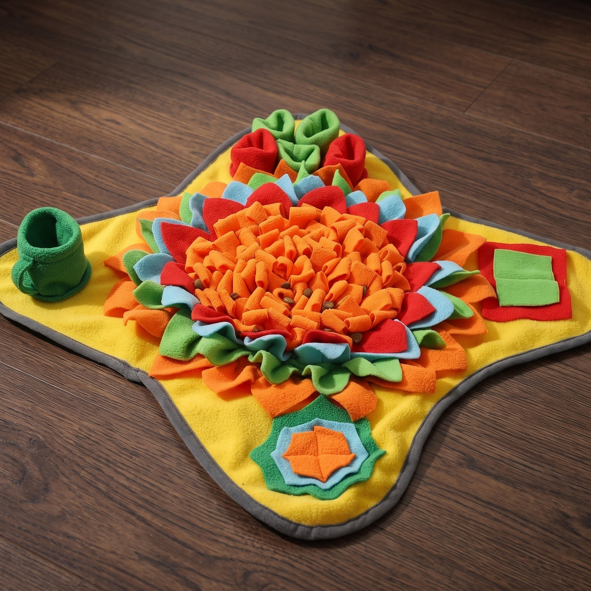 A colorful sensory play mat for toddlers, featuring a yellow base with a gray border and various textured felt tabs in orange, red, green, and blue, displayed on a dark wood floor.